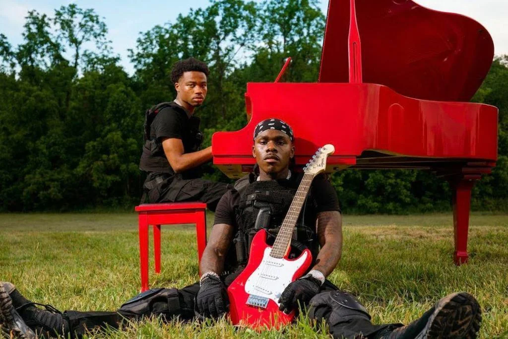 Da Baby and Roddy Rich positioned around a red piano and red electric guitar outdoors